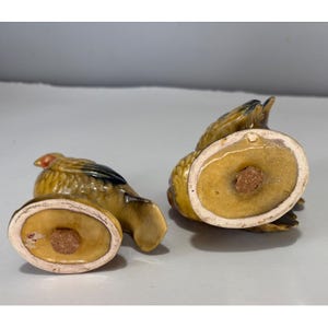 May include: A pair of vintage ceramic bird-shaped salt and pepper shakers. The shakers are primarily yellow with brown and black accents, and feature a red head. The bottom of each shaker has a cork stopper.