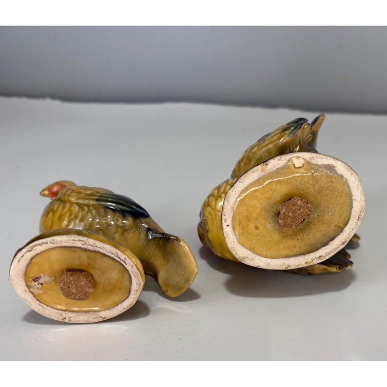 May include: A pair of vintage ceramic bird-shaped salt and pepper shakers. The shakers are primarily yellow with brown and black accents, and feature a red head. The bottom of each shaker has a cork stopper.
