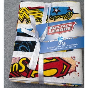 May include: A stack of five Justice League themed cotton fabric pieces, tied with a white ribbon. The fabric features comic book graphics of Wonder Woman and Superman. The tag indicates the fabric is 18 inches by 21 inches.