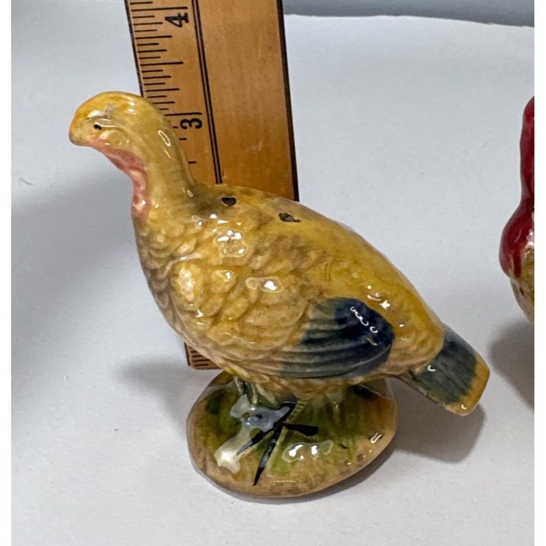 May include: A ceramic turkey figurine with a yellow body, blue wing accents, and a red neck. The turkey is perched on a green base with black accents. The figurine is approximately 3 inches tall.