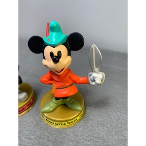 K&ouml;nnte beinhalten: Eine Plastikfigur von Mickey Mouse, verkleidet als der tapfere kleine Schneider, mit einer Schere in der Hand. Die Figur steht auf einem goldenen Sockel mit der Aufschrift "Brave Little Tailor".