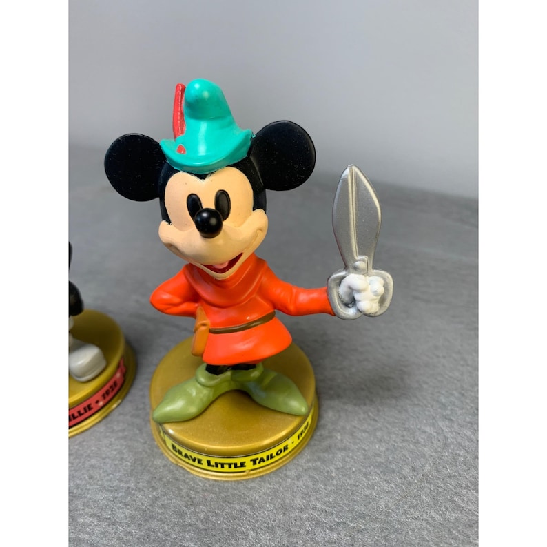 K&ouml;nnte beinhalten: Eine Plastikfigur von Mickey Mouse, verkleidet als der tapfere kleine Schneider, mit einer Schere in der Hand. Die Figur steht auf einem goldenen Sockel mit der Aufschrift "Brave Little Tailor".