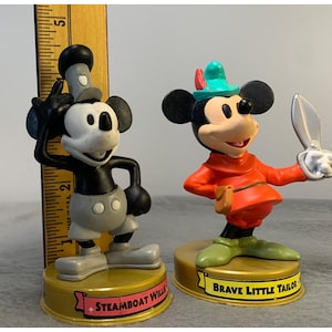 K&ouml;nnte beinhalten: Zwei Vintage-Mickey-Mouse-Figuren. Die Figur links ist schwarz-wei&szlig; mit einem grauen Hut und einer grauen Hose. Die Figur rechts tr&auml;gt ein rotes Hemd, eine gr&uuml;ne Hose und einen gr&uuml;nen Hut mit einer Feder. Die Figur links ist mit "Steamboat Willie" und die Figur rechts mit "Brave Little Tailor" beschriftet.