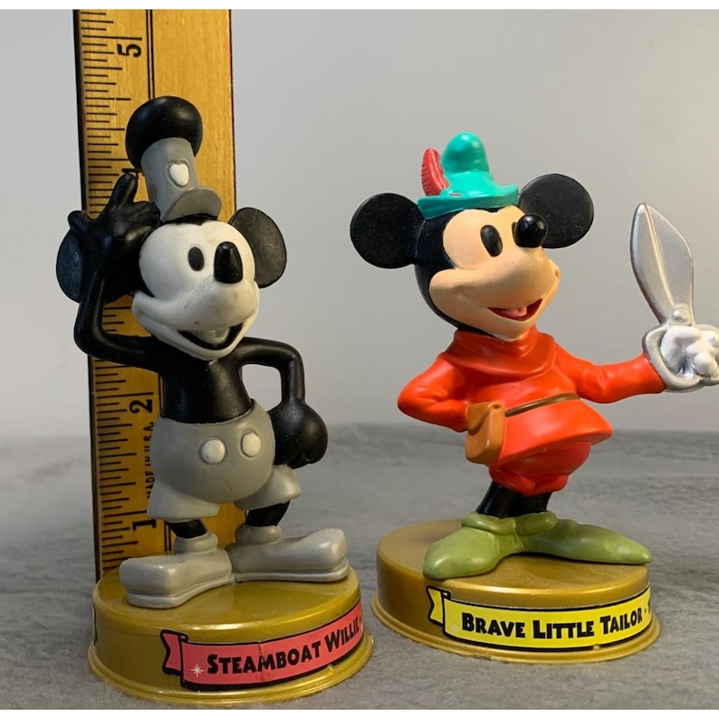 K&ouml;nnte beinhalten: Zwei Vintage-Mickey-Mouse-Figuren. Die Figur links ist schwarz-wei&szlig; mit einem grauen Hut und einer grauen Hose. Die Figur rechts tr&auml;gt ein rotes Hemd, eine gr&uuml;ne Hose und einen gr&uuml;nen Hut mit einer Feder. Die Figur links ist mit "Steamboat Willie" und die Figur rechts mit "Brave Little Tailor" beschriftet.