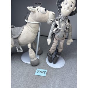 K&ouml;nnte beinhalten: Zwei Pl&uuml;schtiere: ein hellgraues Pferd mit einem grauen Sattel und eine Cowboy-Puppe. Die Cowboy-Puppe tr&auml;gt eine graue Weste, einen gemusterten Schal und einen Cowboyhut. Beide Spielzeuge stehen auf wei&szlig;en St&auml;ndern.