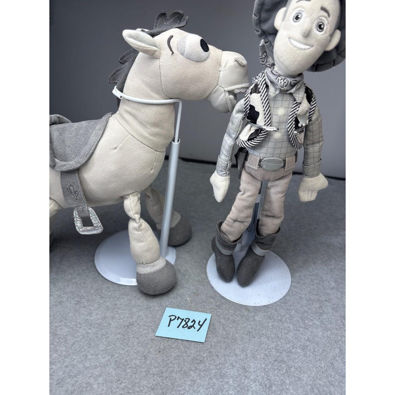 K&ouml;nnte beinhalten: Zwei Pl&uuml;schtiere: ein hellgraues Pferd mit einem grauen Sattel und eine Cowboy-Puppe. Die Cowboy-Puppe tr&auml;gt eine graue Weste, einen gemusterten Schal und einen Cowboyhut. Beide Spielzeuge stehen auf wei&szlig;en St&auml;ndern.