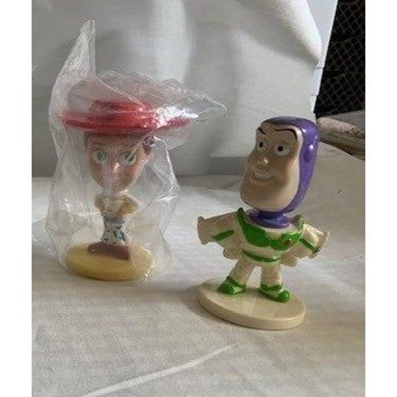 May include: Two Toy Story bobblehead figures: Jessie in a red hat and Buzz Lightyear in a green and white spacesuit. Both figures are on oval bases and appear to be made of plastic.