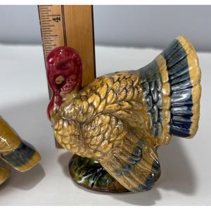 May include: A ceramic turkey figurine with a red head and wattle, yellow and brown body, and blue and yellow tail feathers. The figurine is on a green base and appears to be a salt or pepper shaker. A wooden ruler is in the background.