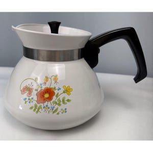 May include: A white teapot with a black handle and lid, featuring a floral pattern of orange, yellow, and blue flowers with green leaves. A silver-colored metal band encircles the upper portion of the teapot body, separating the lid from the base.