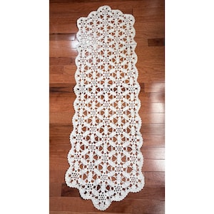 May include: A cream-colored, open-weave crochet table runner. The runner has a scalloped edge and a repeating geometric pattern. The runner is displayed on a wooden surface.