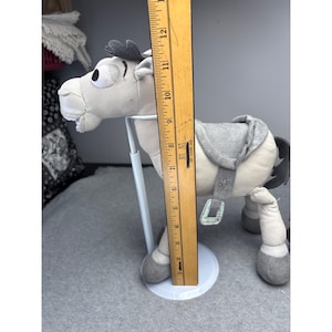 K&ouml;nnte beinhalten: Ein Pl&uuml;schpferd in Grau- und Wei&szlig;t&ouml;nen mit Sattel und dekorativen Akzenten. Das Spielzeug ist etwa 23 cm hoch, gemessen mit einem Holzlineal. Das Pferd hat ein skurriles Design.
