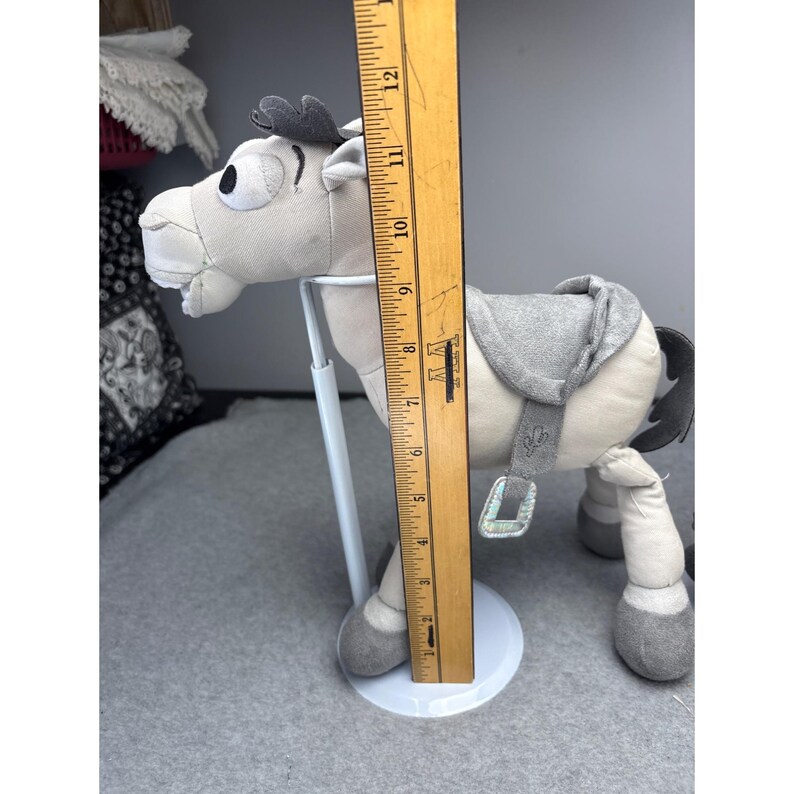 K&ouml;nnte beinhalten: Ein Pl&uuml;schpferd in Grau- und Wei&szlig;t&ouml;nen mit Sattel und dekorativen Akzenten. Das Spielzeug ist etwa 23 cm hoch, gemessen mit einem Holzlineal. Das Pferd hat ein skurriles Design.