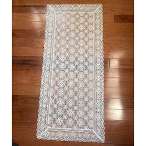 May include: A rectangular, white lace table runner with a detailed floral pattern. The runner has a scalloped edge and is laid out on a wooden surface. The lace design is intricate and delicate, adding a touch of elegance.