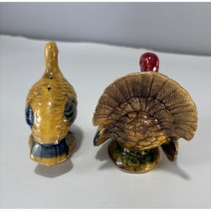 May include: A pair of ceramic turkey salt and pepper shakers. One shaker is facing forward with its tail feathers fanned out, and the other is facing away. The turkeys are primarily yellow, brown, and blue with red accents.
