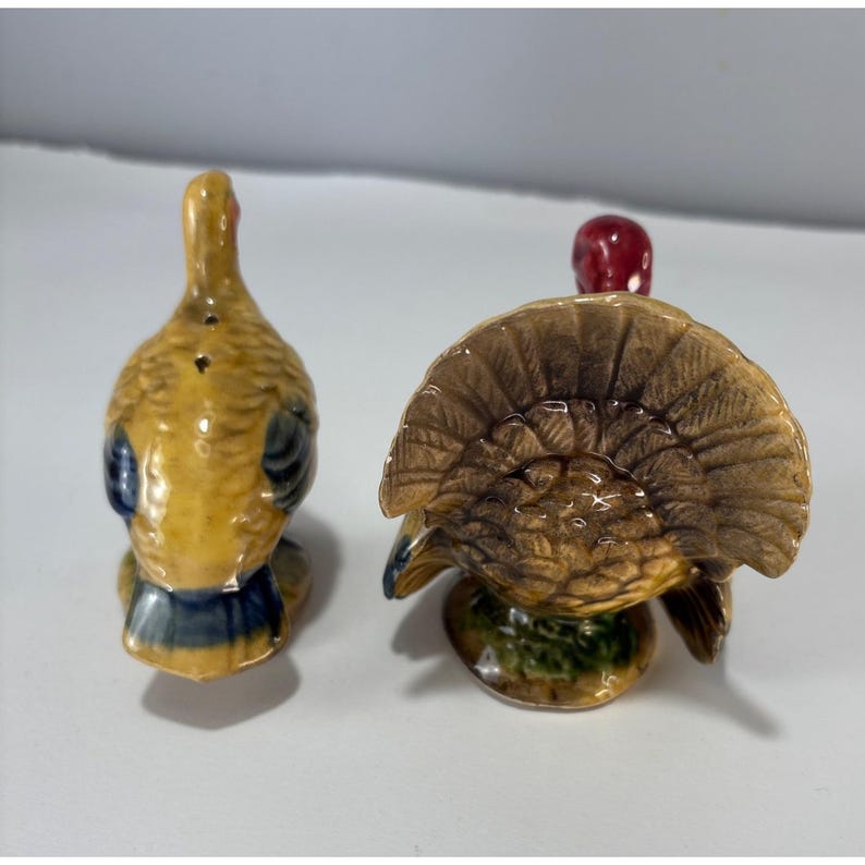 May include: A pair of ceramic turkey salt and pepper shakers. One shaker is facing forward with its tail feathers fanned out, and the other is facing away. The turkeys are primarily yellow, brown, and blue with red accents.