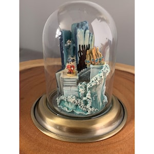 May include: A glass dome with a brass base encloses a miniature scene featuring Mickey Mouse standing on a staircase in front of a blue and white building. The scene is set against a backdrop of blue water with white foam.
