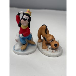 May include: Two ceramic figurines featuring Goofy and Pluto. Goofy wears a red sweater, blue pants, and a yellow hat while ice skating on a white base. Pluto is a tan dog with a black collar, crouching on a white base while chewing a bone.