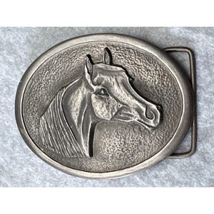 May include: Oval-shaped silver-toned metal belt buckle featuring a detailed relief of a horse's head. The horse's profile is centered within a textured border, with a rectangular bar on the right side for belt attachment. The buckle has a vintage or antique appearance.