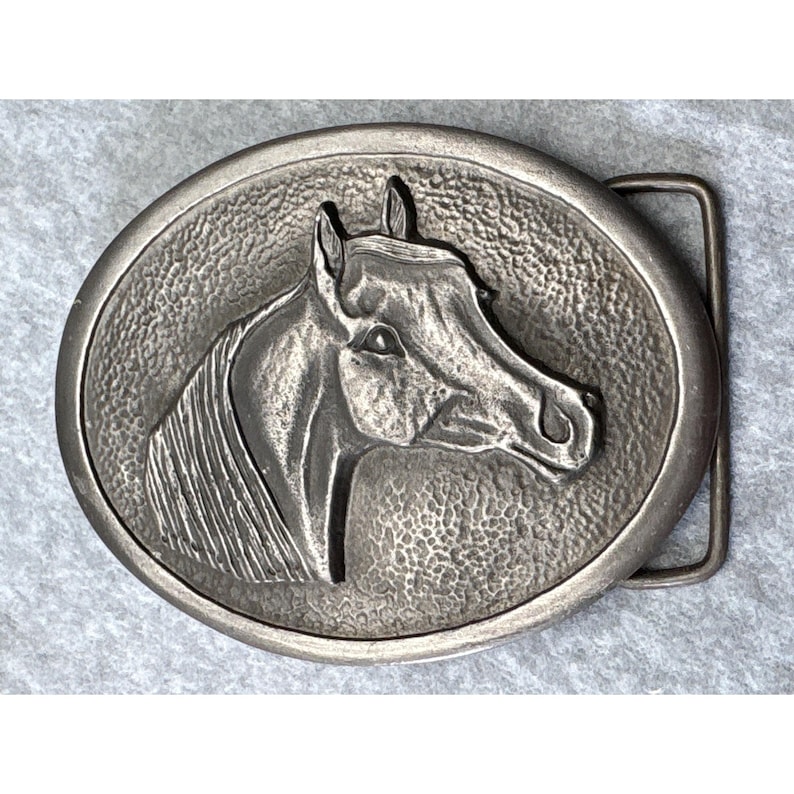 May include: Oval-shaped silver-toned metal belt buckle featuring a detailed relief of a horse's head. The horse's profile is centered within a textured border, with a rectangular bar on the right side for belt attachment. The buckle has a vintage or antique appearance.
