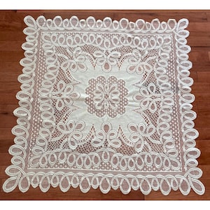 May include: A square off-white lace doily featuring a central floral motif surrounded by intricate scrollwork patterns. The border is defined by a continuous series of looped, rounded shapes, creating a decorative edge around the entire piece.