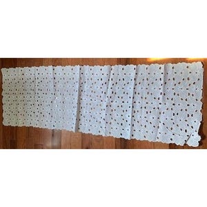 May include: White, openwork table runner with a floral design. The runner is rectangular with scalloped edges and a repeating pattern of cut-out flower shapes. The runner is displayed on a wooden surface.
