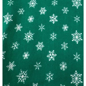 Op de afbeelding: Een diepgroene stof met een herhalend patroon van witte sneeuwvlokken in verschillende groottes en complexe geometrische ontwerpen. Het oppervlak heeft een zichtbare geweven textuur en de sneeuwvlokken zijn gelijkmatig over het materiaal verdeeld.