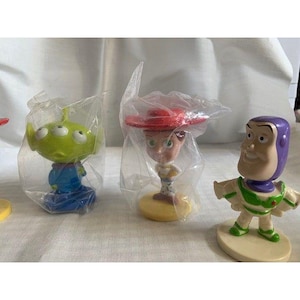 May include: A collection of Toy Story character figurines, including an alien, Jessie, and Buzz Lightyear. The alien is green with three eyes, the Jessie figurine has a red hat, and Buzz Lightyear is in a white and green spacesuit.