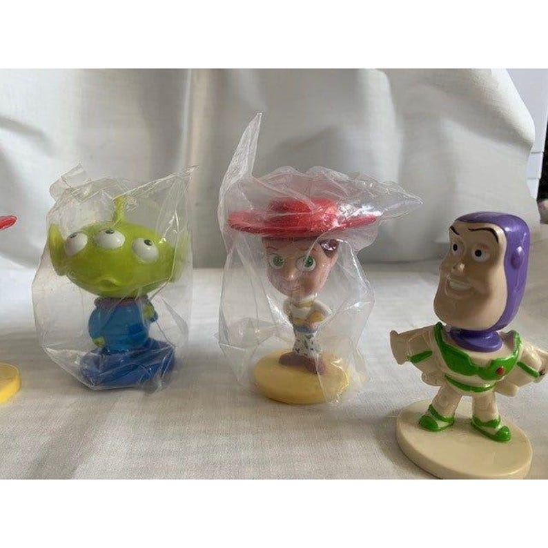 May include: A collection of Toy Story character figurines, including an alien, Jessie, and Buzz Lightyear. The alien is green with three eyes, the Jessie figurine has a red hat, and Buzz Lightyear is in a white and green spacesuit.