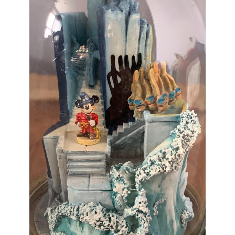 May include: A glass snow globe with a blue and white castle scene. Mickey Mouse is standing in front of the castle, wearing a red and gold outfit. The snow globe is filled with white glitter.