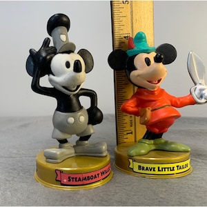 K&ouml;nnte beinhalten: Zwei Vintage-Mickey-Mouse-Figuren. Die Figur links ist schwarzwei&szlig; und tr&auml;gt die Aufschrift "Steamboat Willie". Die Figur rechts ist farbig und tr&auml;gt die Aufschrift "Brave Little Tailor".
