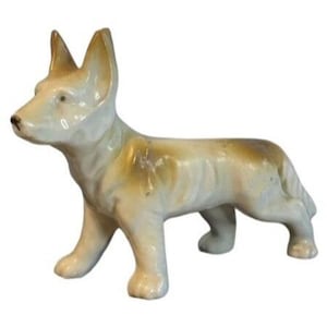 May include: A ceramic figurine of a dog in a standing pose. The dog has a light beige coat with brown markings on its back. The ears are upright, and the dog appears to be a German Shepherd. The figurine is a decorative item.