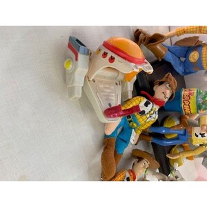 May include: A collection of Toy Story character plush toys, including Woody, Buzz Lightyear, and others. Woody is wearing a cowboy hat, vest, and blue jeans. Buzz Lightyear has a white and gray space suit with an orange helmet. The toys are on a white surface.