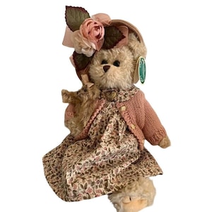 May include: A plush teddy bear with light brown fur wearing a pink knit cardigan over a floral print dress. The bear has a hat adorned with pink and white fabric flowers and holds a smaller teddy bear. A green tag is attached to the bear's ear.