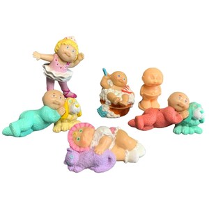 May include: A collection of six small plastic baby figurines. One wears a pink tutu, one sits in a bubble bath, one stands in a diaper, two lie down hugging small animal figures, and one sleeps on a purple cat. The figures feature various colors.