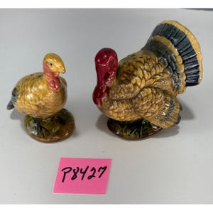 May include: A pair of ceramic turkey figurines, likely salt and pepper shakers. The turkeys are painted with brown, yellow, red, and blue hues. One turkey is standing upright, and the other is in a seated position. The base of each turkey is green.