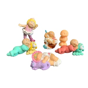 May include: A collection of vintage Cabbage Patch Kids figurines. The figurines depict babies in various poses, including a ballerina, a baby in a tub, and babies with animal companions. The color palette includes pastels and bright colors.