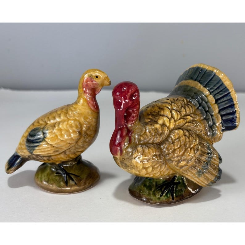 May include: A pair of ceramic turkey figurines. One is a hen, the other a tom. The hen is yellow with blue and black accents. The tom has a red wattle and a fan of blue, yellow, and black feathers. Both are on green bases.