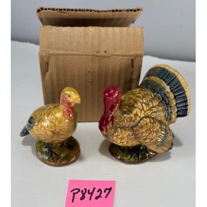 May include: A pair of ceramic turkey figurines, one standing and one sitting, with yellow, red, and blue coloring. The figurines are on a green base. A cardboard box is in the background. A pink sticker with the text "P8427" is in the foreground.