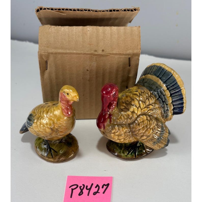May include: A pair of ceramic turkey figurines, one standing and one sitting, with yellow, red, and blue coloring. The figurines are on a green base. A cardboard box is in the background. A pink sticker with the text "P8427" is in the foreground.
