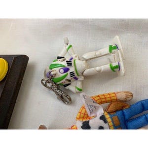 May include: A white and green Buzz Lightyear keychain toy with purple accents. The toy is lying on a white surface next to a Woody doll. The keychain is attached to a silver metal clasp.