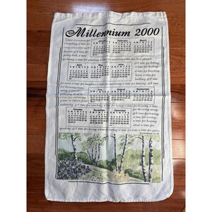 Könnte beinhalten: Off-white Millennium 2000 Kalender-Geschirrtuch mit einer Landschaftsszene am unteren Rand. Der Kalender enthält Monate, Daten und Text aus Prediger 3:1-8. Das Geschirrtuch ist rechteckig und aus weichem Stoff.