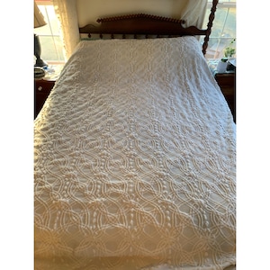 May include: Cream-colored, textured bedspread with a repeating circular pattern. The bedspread is draped over a dark wooden bed frame. The room has a window with natural light and a bedside table with a lamp.