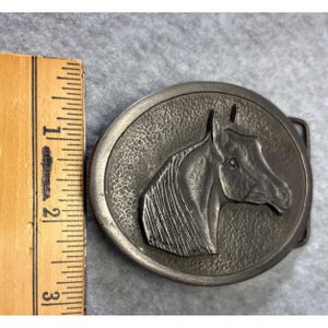 May include: Oval-shaped, silver-toned belt buckle featuring a detailed relief of a horse's head. The horse's head is centered within a textured circle. The buckle measures approximately 3 inches in length.