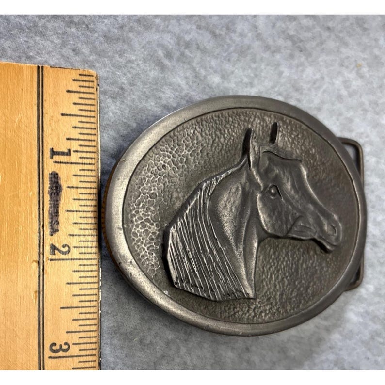 May include: Oval-shaped, silver-toned belt buckle featuring a detailed relief of a horse's head. The horse's head is centered within a textured circle. The buckle measures approximately 3 inches in length.
