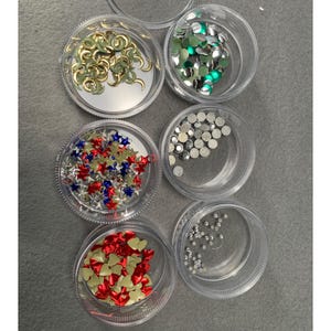 May include: Six clear circular containers filled with various craft embellishments. The collection includes gold crescent moons, green circular discs, red, white, and blue stars, silver circular discs, red and cream hearts, and tiny clear rhinestones.