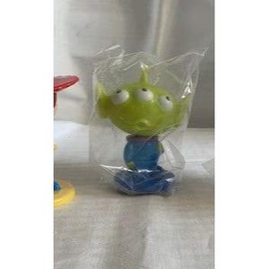 May include: A green, three-eyed alien figure from the Toy Story franchise, packaged in a clear plastic bag. The figure has a blue body and is standing on a blue base. The alien's head is a lighter shade of green than its body.