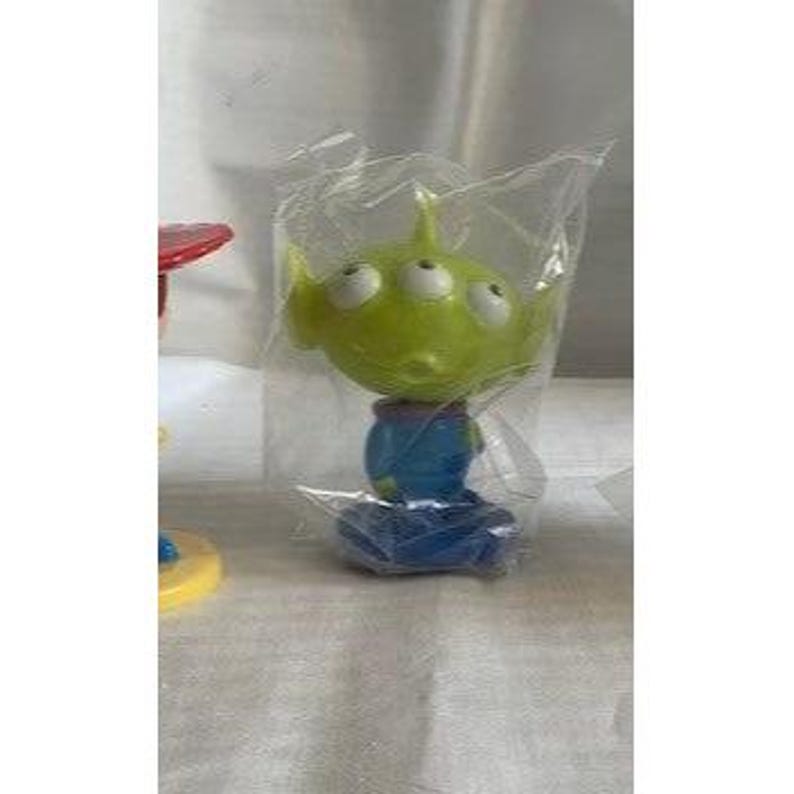 May include: A green, three-eyed alien figure from the Toy Story franchise, packaged in a clear plastic bag. The figure has a blue body and is standing on a blue base. The alien's head is a lighter shade of green than its body.