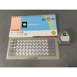 May include: A Cricut Damask Décor set, featuring a keyboard with decorative keys, a design book, and a gray cartridge. The design book has a blue background with floral designs and the Cricut logo. The keyboard has the text "Damask Décor".