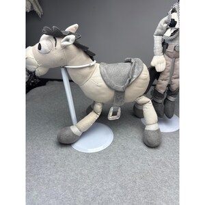 K&ouml;nnte beinhalten: Ein Pl&uuml;schpferd mit einem grauen Sattel und M&auml;hne. Das Pferd ist hellbraun mit grauen Hufen und einem wei&szlig;en Zaumzeug. Es steht auf einem wei&szlig;en Sockel. Eine weitere Spielfigur ist im Hintergrund teilweise sichtbar.