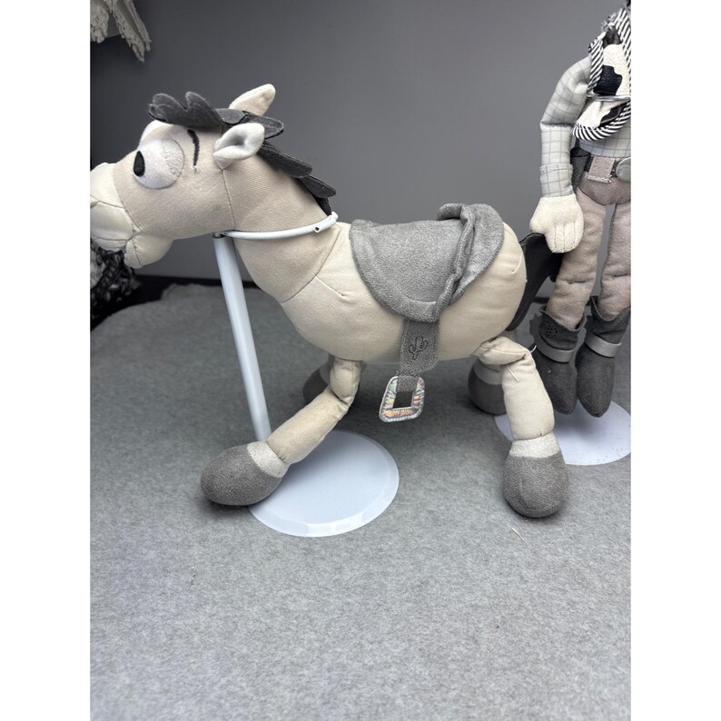 K&ouml;nnte beinhalten: Ein Pl&uuml;schpferd mit einem grauen Sattel und M&auml;hne. Das Pferd ist hellbraun mit grauen Hufen und einem wei&szlig;en Zaumzeug. Es steht auf einem wei&szlig;en Sockel. Eine weitere Spielfigur ist im Hintergrund teilweise sichtbar.
